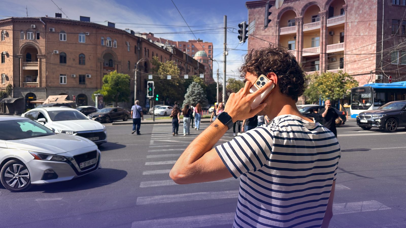 A man on a phone call at a busy city crossing with cars and pedestrians behind him