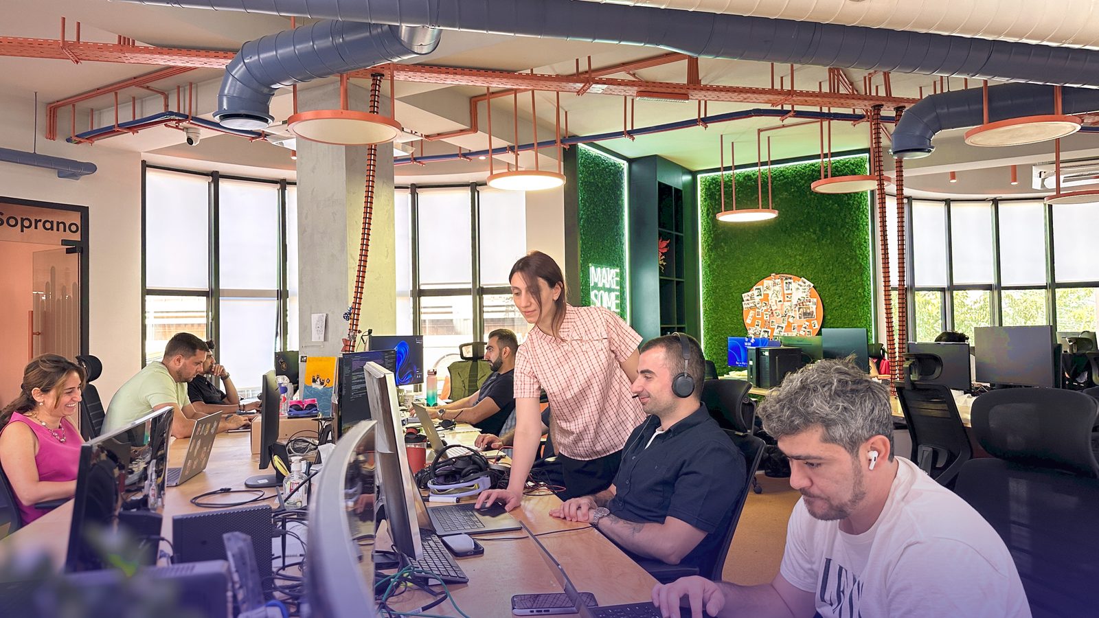 An open-plan office with several people at desks, one standing and talking to a colleague wearing a headset
